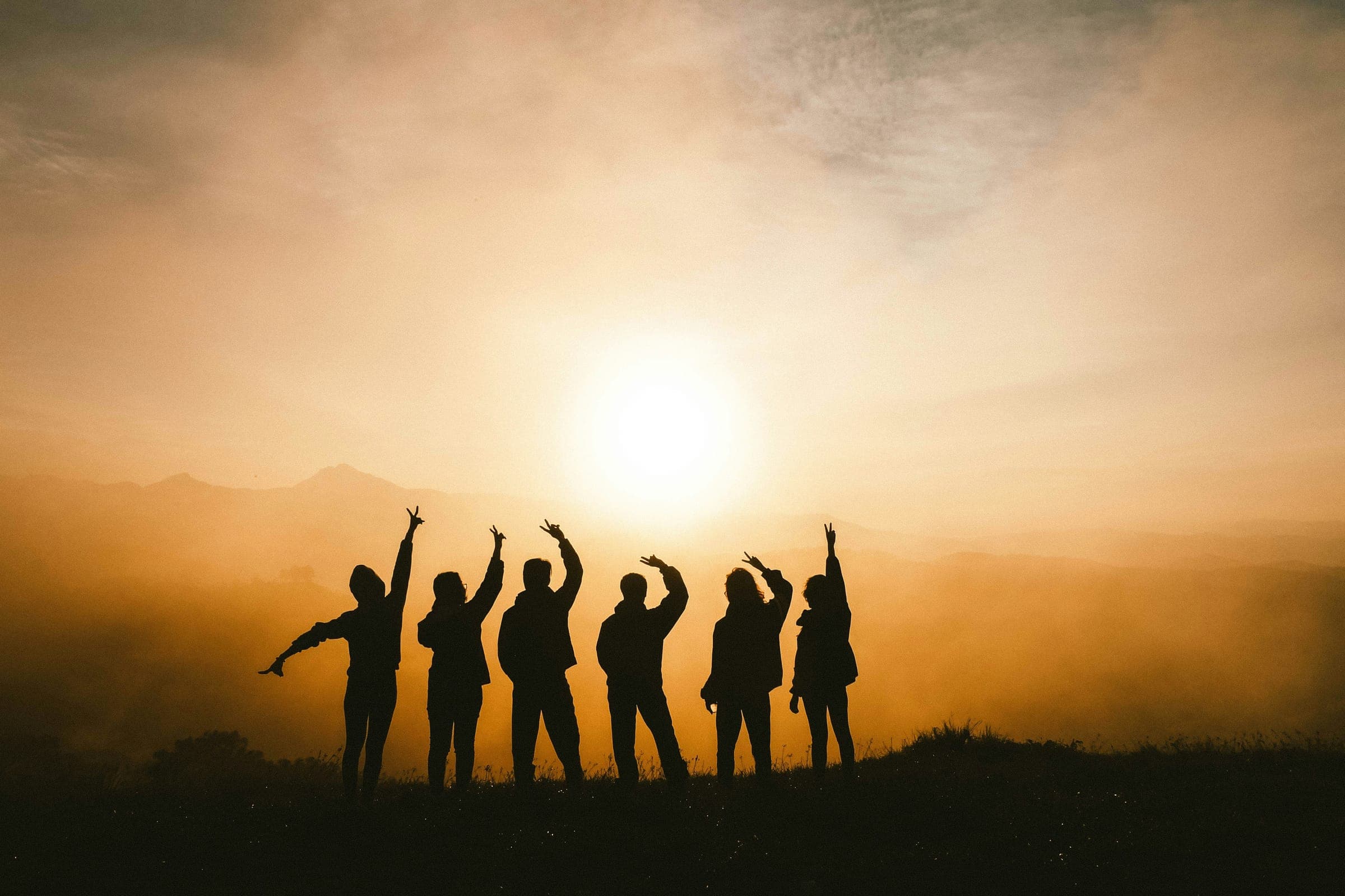A group of friends standing together on a hilltop at sunset, arms raised